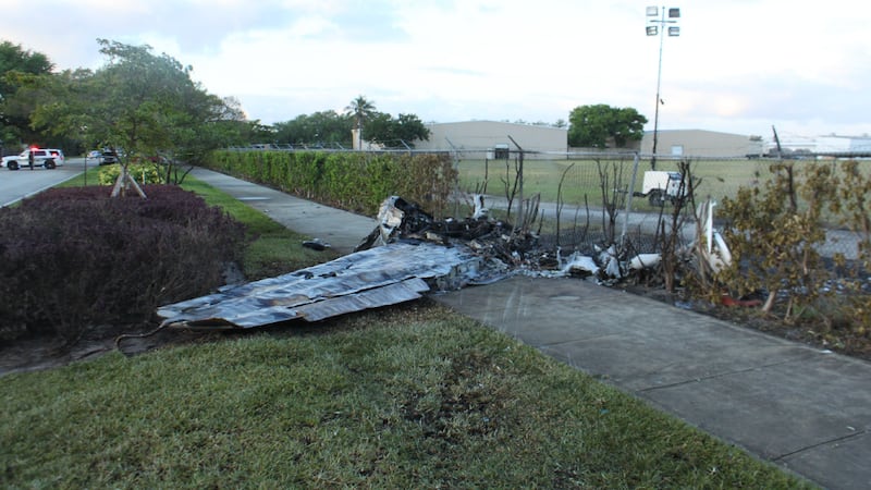 Gouge marks in the grash are seen in the foreground, mangled metal and a damaged fence are...